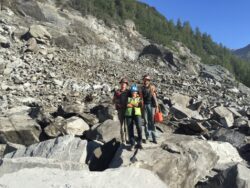 Three individuals wearing hard hats and backpacks are standing on a rocky terrain with a large pile of boulders in the background, suggesting they may be geologists or hikers exploring a rugged area.