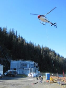 A helicopter is seen hoisting cargo near an industrial facility with forested hills in the background, while personnel on the ground oversee the operation.