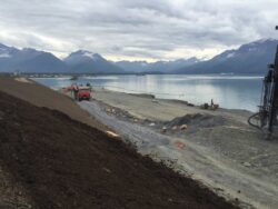 Construction equipment and workers on a sloped terrain with a body of water and mountains in the background.