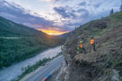 Workers on a steep hillside at dusk with safety gear, overlooking a river and road as the sun sets in the distance.