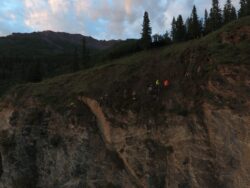 A group of people wearing high-visibility vests are traversing through a rugged terrain on a hillside during dusk.