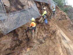 Workers in safety gear installing protective netting on a steep rock face.