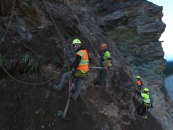 Construction or maintenance workers in high-visibility vests with safety helmets equipped with headlamps are secured with harnesses while working on a steep rock face beside a road.