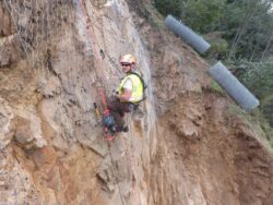 A worker wearing a safety harness is installing wire mesh on a steep slope to prevent rockslides.