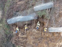 Workers in safety gear are installing wire mesh on a sloped terrain to prevent erosion or landslides.