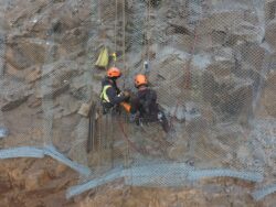 Two workers in safety harnesses are performing maintenance or construction work on a steep rock face, secured with protective netting.