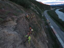 Workers in high visibility gear secure a slope beside a road near a river during dusk.