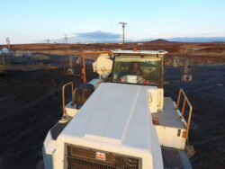 A worker is smiling from the driver's seat of a large industrial vehicle on a dirt terrain with an open landscape and sunset in the background.