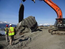 Construction workers inspecting large geotextile tubes being lifted by an excavator at a coastal construction site, with mountains and water visible in the background.