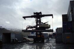A container straddle carrier is operating in an industrial port area, moving a cargo container on a cloudy day with snow-covered hills in the background.