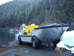 A person standing in front of a large amphibious vehicle by a snowy lakeshore.