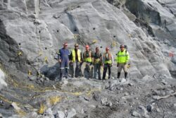 A group of six construction workers wearing hard hats, high visibility vests, and work gear standing in front of a rocky excavation site with marked areas and loose cables on the ground.