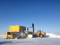 Forklift operating near a temporary modular structure and shipping containers on a snow-covered expanse under a clear blue sky.