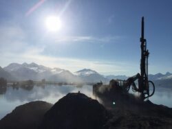 Drilling rig operating on a clear day with a mountainous backdrop and a body of water in the foreground.