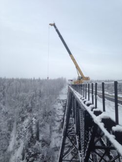 A crane is positioned on a snow-covered bridge, with its boom extended out over the side, amidst a wintery forest landscape.