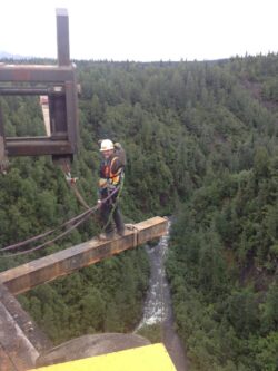 A construction worker in safety gear is standing on a narrow beam above a river, secured with safety lines, possibly engaged in bridge inspection or maintenance.