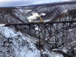 A crane mounted on a railroad bridge extending over a snowy ravine, with forested slopes and a river visible in the background.