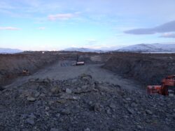 A construction or mining site with machinery and piled up earth, set against a backdrop of distant mountains and a cloudy sky at dusk.