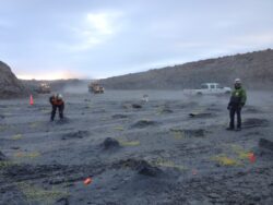 Workers at a mining or construction site with vehicles in the background, surrounded by a dusty environment at dusk or dawn.
