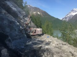 An excavator perched on a steep, rocky incline is in the process of either drilling or excavation work, set against a backdrop of mountains and a body of water.