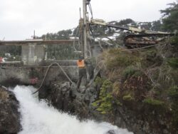 A worker in reflective safety gear is overseeing a drilling operation near a rushing water channel, with a large drill rig stationed above the rocky terrain.