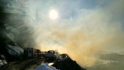 Sunlight filters through mist, casting rays over a snowy mountain road with parked vehicles and containers.