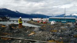 A construction worker in a high-visibility vest and hard hat stands facing a harbor with mountains in the distance, amidst a landscape of rocks and debris indicative of construction or excavation work.