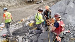 Workers in safety gear, including helmets and high-visibility vests, are engaged in work at a mining site with boxes and tools on the ground around them.