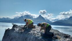 Two workers in high visibility vests and safety helmets are securing ropes on a rocky outcrop with a scenic backdrop of a lake and mountains under a clear blue sky.