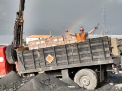 A construction worker in an orange vest and hard hat stands with outstretched arms on the back of a dump truck, with a rainbow visible in the cloudy sky behind him.