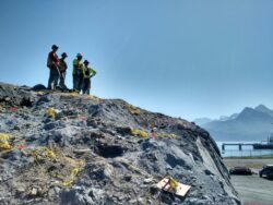 Four construction workers wearing hard hats stand atop a rocky mound with surveying equipment, overlooking a body of water with mountains in the distance.