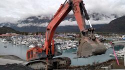 An orange excavator with its bucket raised overlooks a marina filled with boats, with mountains and clouds in the background.