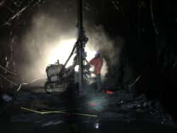 A miner operates a drill machine inside a dimly lit underground mine tunnel, surrounded by rock walls and illuminated by artificial lighting.