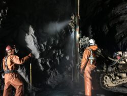 Two miners in protective gear working with heavy machinery in an underground mine tunnel.