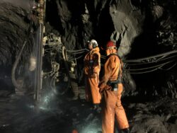 Two miners in orange safety suits and helmets are standing inside a dark mine, illuminated by artificial light, working near a drill machine with cables running along the rocky surface.