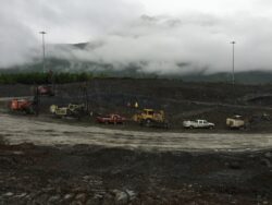 Construction machinery, including excavators and trucks, on a muddy worksite with fog-shrouded mountains in the background.