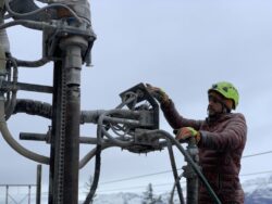 A worker in a helmet and safety gear operates heavy machinery outdoors with snowy mountains in the background.