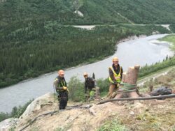 Three individuals wearing hard hats and high-visibility vests stand on a forested slope above a winding river, with one person holding a rope tied to a piece of heavy, rusted equipment.