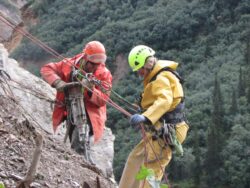 Two climbers wearing helmets and safety gear are preparing or adjusting ropes on a rocky slope with green vegetation in the background.