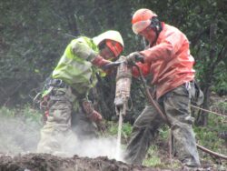 Two workers in safety gear operating a heavy-duty drill outdoors.