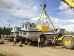 A heavy-duty crane is lifting a large, silver, amphibious vehicle onto a flat surface while several workers in high-visibility vests oversee the operation at an outdoor location with trees in the background.