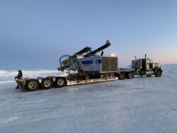 A person stands next to a large truck hauling heavy machinery on a sled across a snowy landscape during twilight.