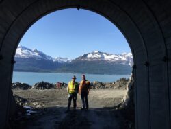 Two workers in high-visibility vests stand at the entrance of a tunnel with a scenic backdrop of a lake and mountains.