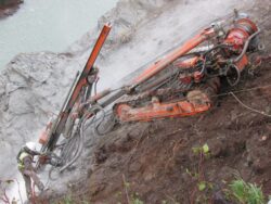 A worker standing beside a large, angled drilling machine at a rocky excavation site.