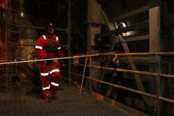 A worker in red and white protective gear is standing inside an industrial facility holding a rope.
