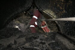 A miner wearing protective gear is working in a cramped and poorly lit underground tunnel.