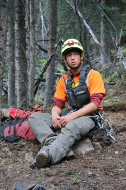 A person sits on a rock in a forest setting, wearing a helmet with a headlamp, an orange vest over a red shirt, and equipped with climbing gear, looking slightly off-camera.