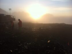 Workers in reflective vests at a construction site at dawn with mountains in the background and fog in the air.