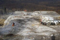 An active construction site with an excavator, a truck, and a worker holding a hose, surrounded by rocky terrain and piles of material.
