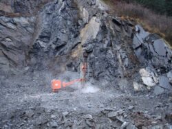 An orange hydraulic rock breaker is being used to break apart a section of rock at a rocky excavation site.